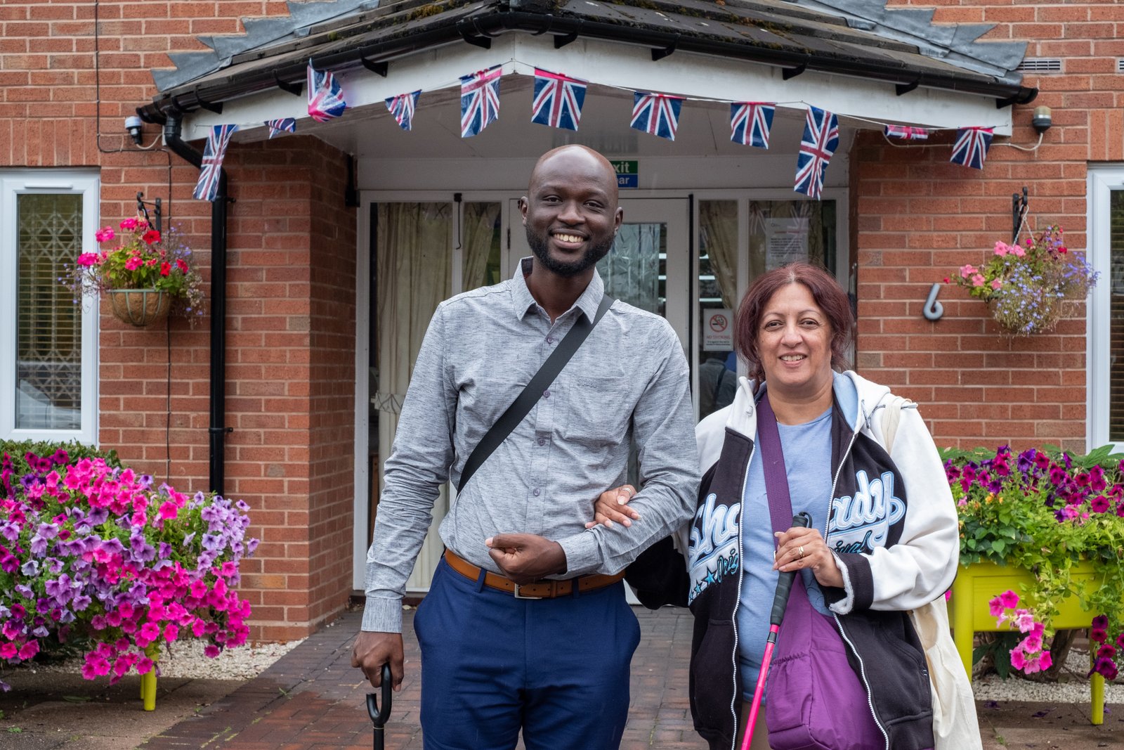 A middle-aged British Asian woman has linked arms with her personal assistant, a young Ghanian man. They are in front of a redbrick building with flower boxes and hanging baskets full of petunias.