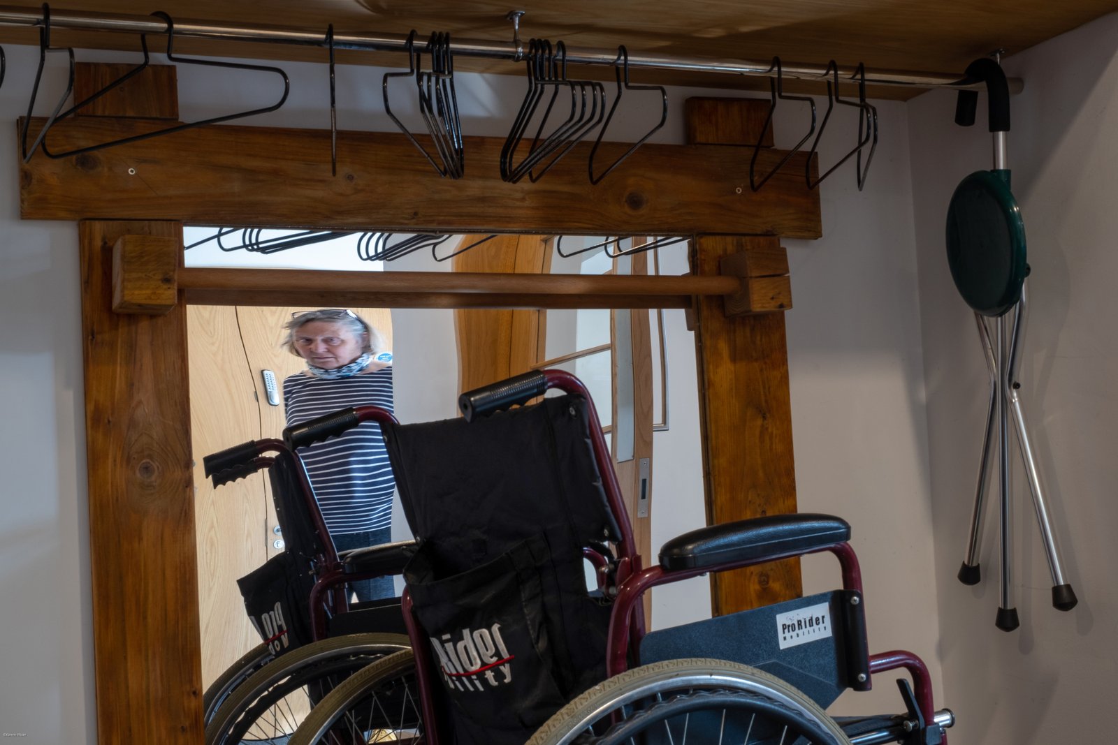 A wheelchair and a folding stool with coat hangers on a clothes rail are reflected in a large mirror, behind which is a distorted reflection of a white woman.