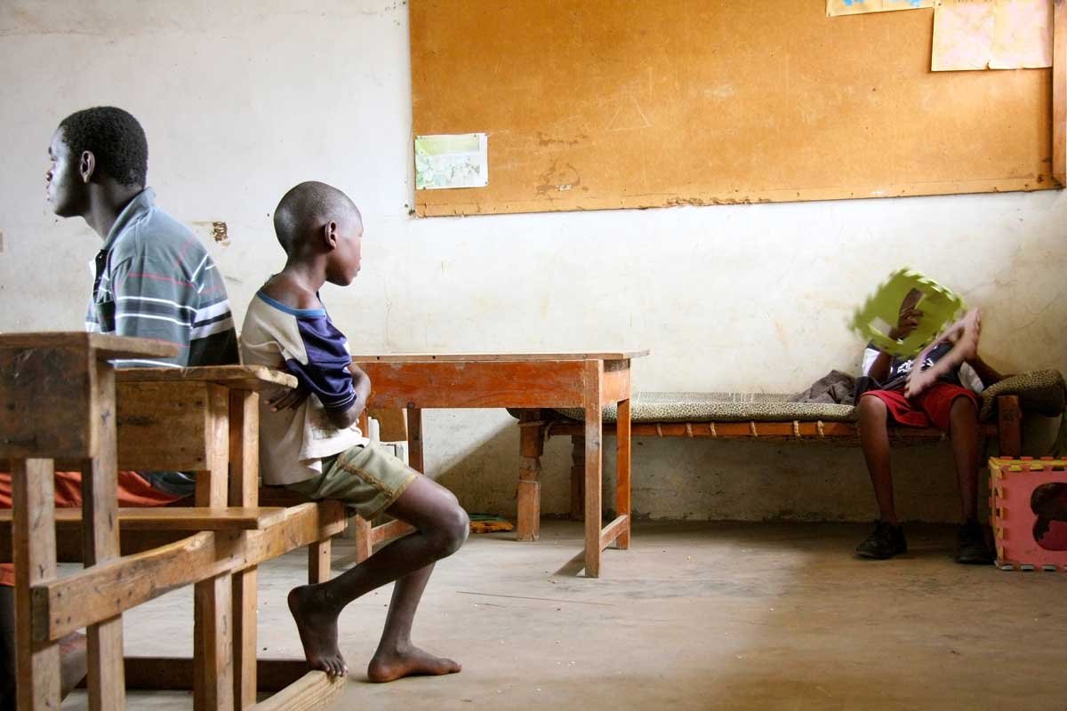 Two boys sit back-to-back in a sparse classroom. A third child reclines on a bed in the corner.