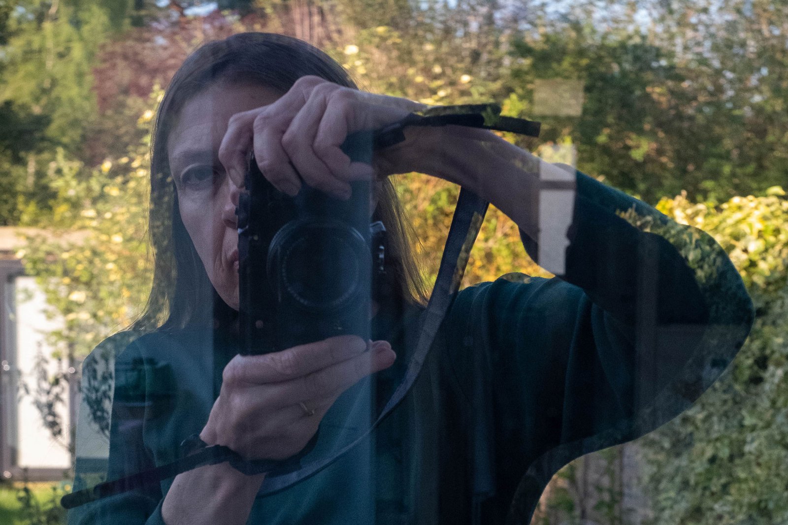 Karren Visser in green long-sleeve T-shirt reflected in window holding camera close so that one half of her face is concealed. A brightly lit garden is behind her.