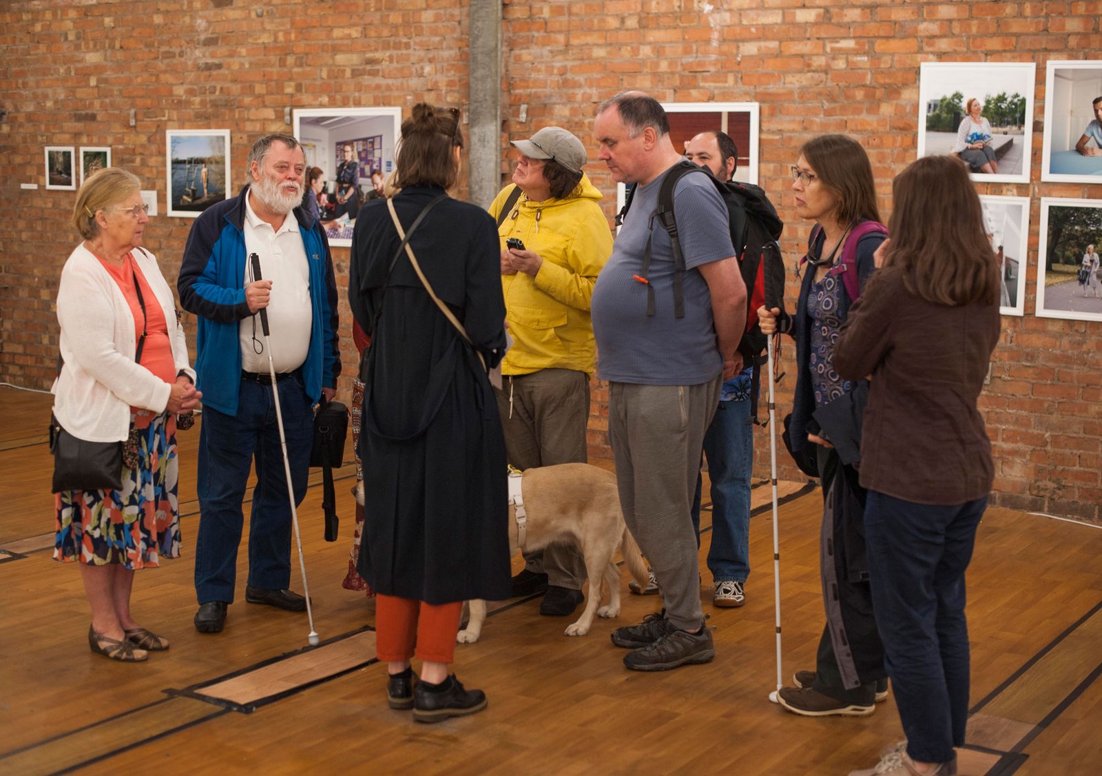 A group of people, two with canes, one with a guide dog, stand in front of a wall of photographs.