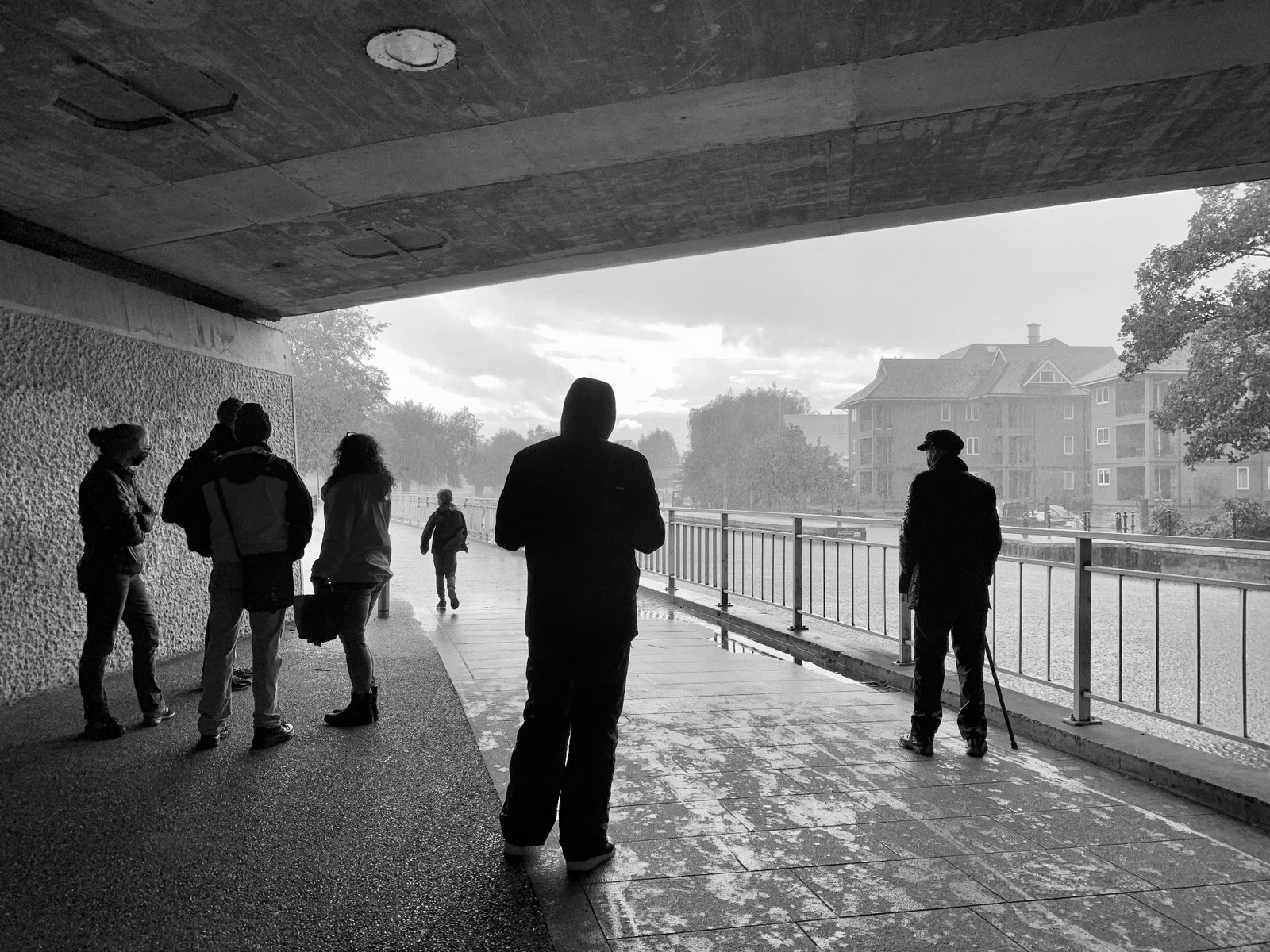 A black and white photograph of pedestrians under a bridge waiting for rain to stop.