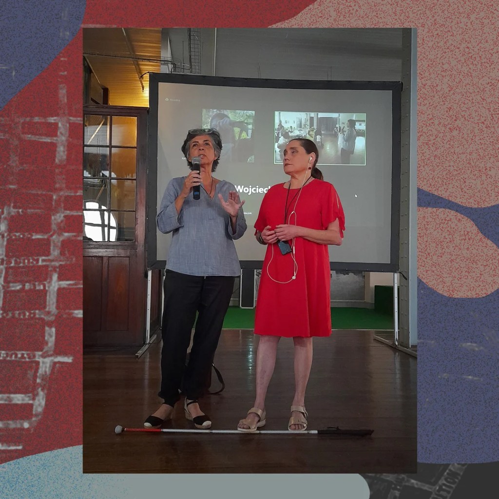 A blind and sighted woman in front of a projector screen showing attendees accessing the event via Zoom speak to an audience not included in the photograph.