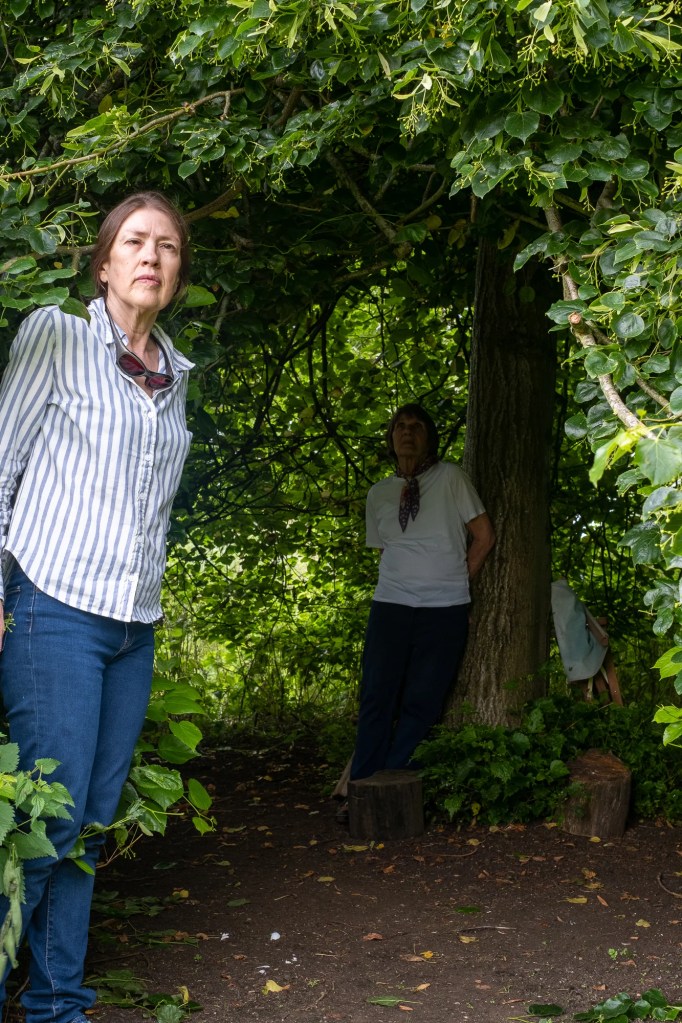 Woman in her fifties stands in the foreground to the left of a leafy tree canopy. Another woman in her eighties is in the background leaning against the tree trunk.