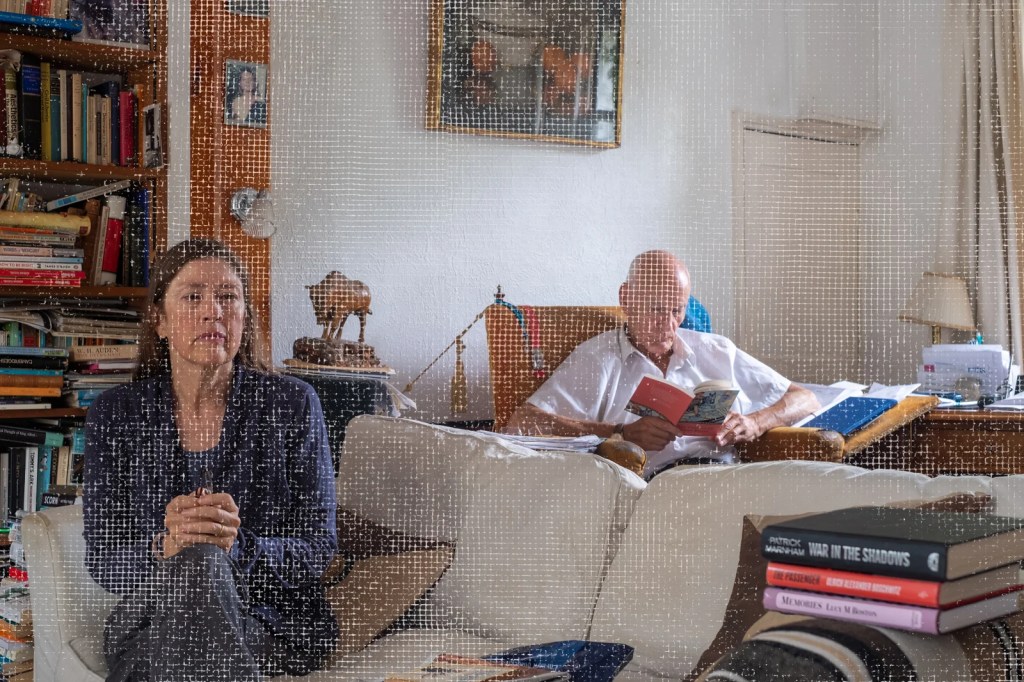 Woman in her fifties is seated on a sofa in the foreground, behind her is a man in his seventies reading. The sitting room is well lit and full of books.