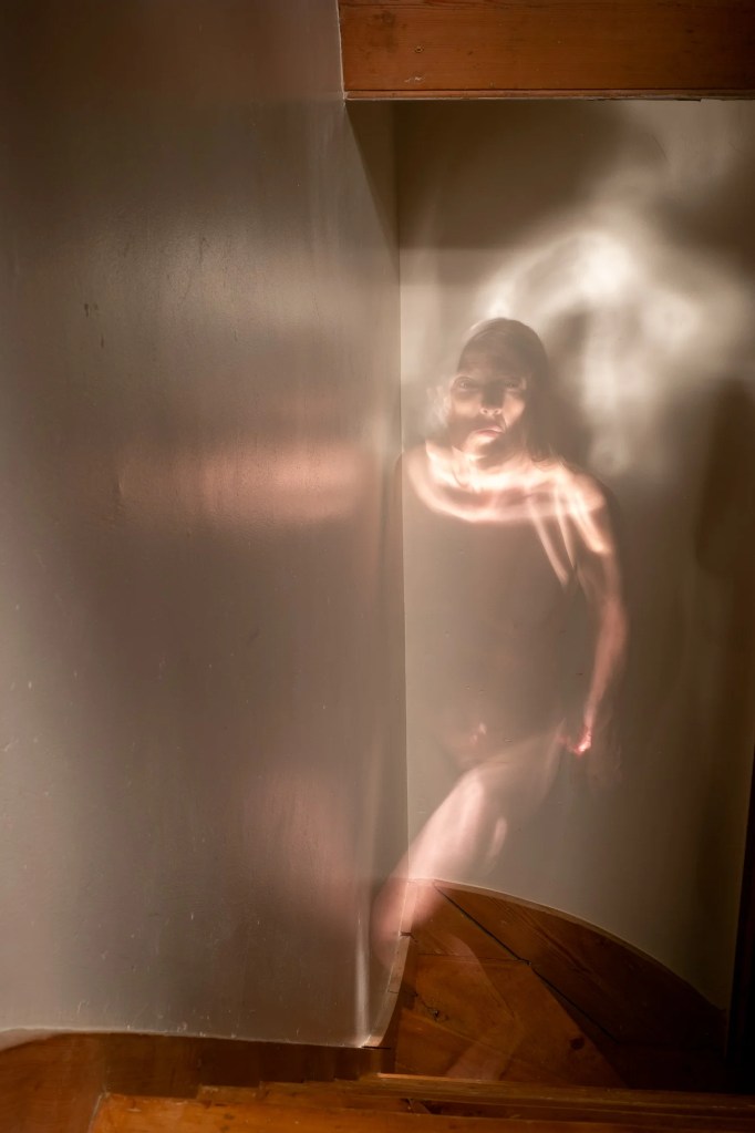 A naked woman half in shadow looks pensive as she walks up a narrow flight of wooden stairs. 
