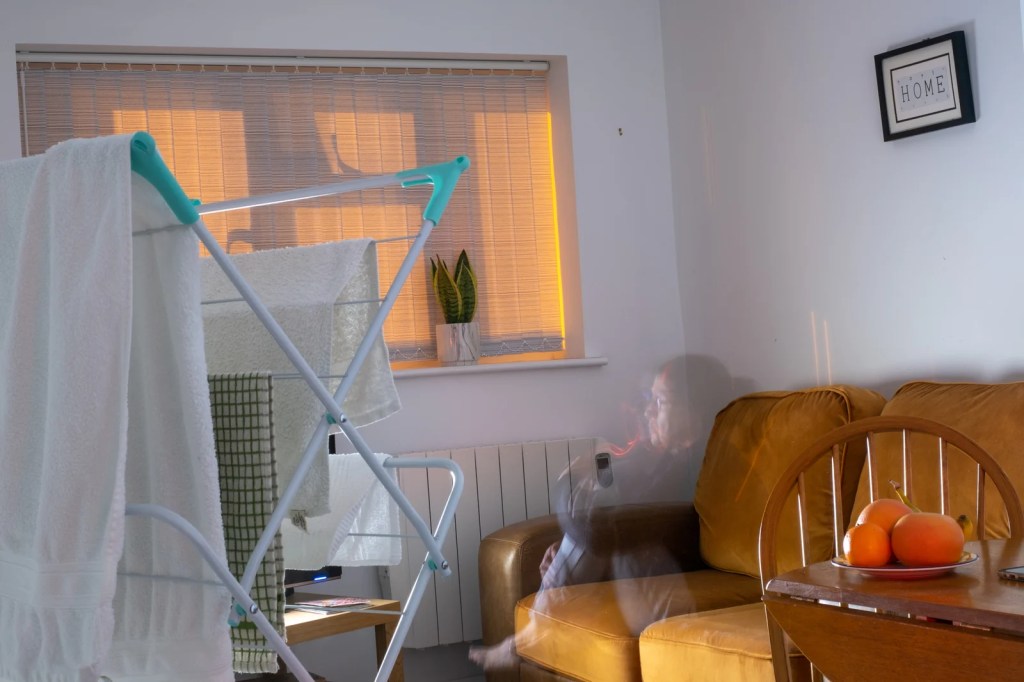 An image of a woman is superimposed seated on a sofa in a cluttered room with a clothes airer or drying rack in the foreground.