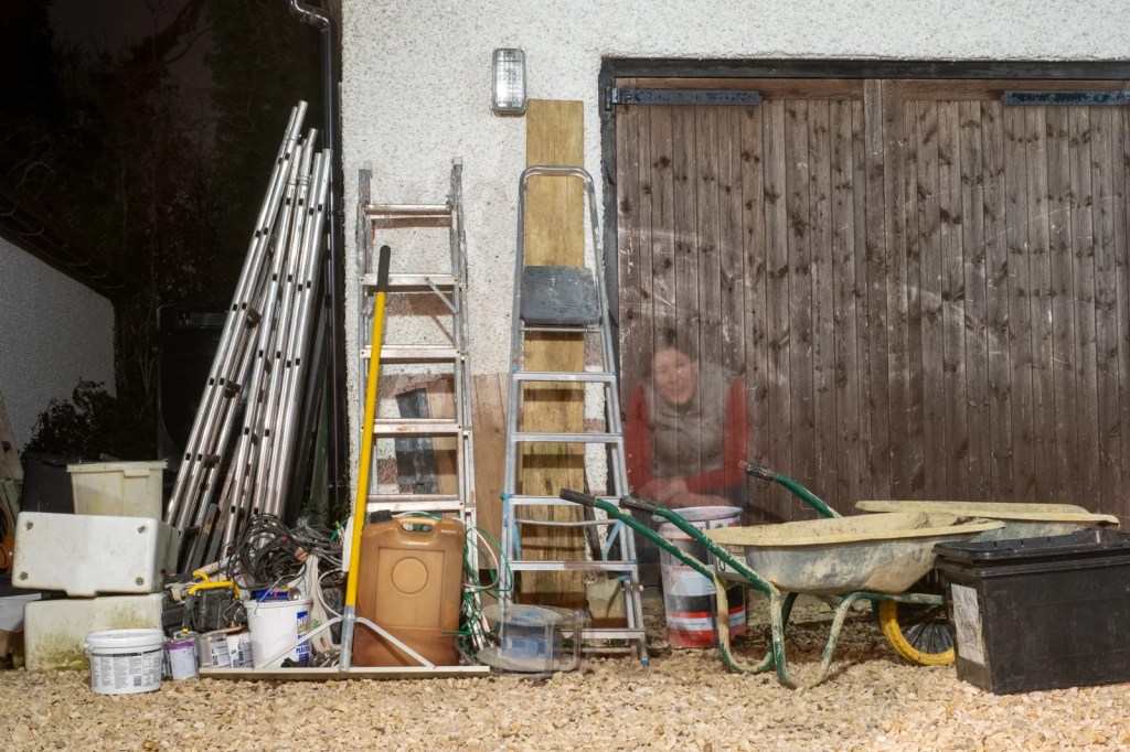 Lit by a security light at night, a woman is seated in front of a workshop door surrounded by building construction materials.