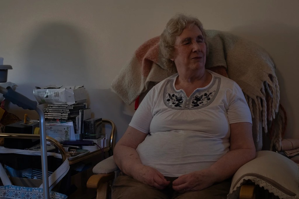 A white woman seated in a comfortable chair in her dimly lit sitting room.