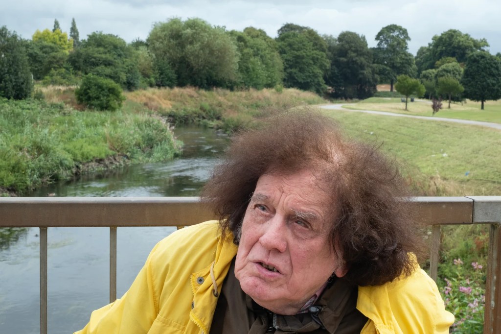 The image on the right: he is in the foreground with a bridge over a stream and trees behind.