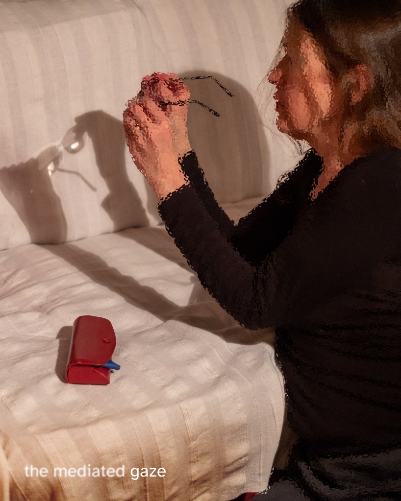 Karren Visser, with long brown hair, kneels beside a light-coloured couch, shown in side profile. She wears a black long-sleeved top and holds a pair of eyeglasses close to her face. Strong light from the left casts a sharp shadow of her hands and the glasses onto the couch. A small red case rests on the couch in front of her. Text in the bottom-left corner reads: “the mediated gaze”. © Karren Visser. Sight Lines, funded by Arts Council England, 2021–22.